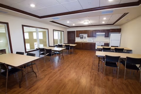 A spacious room with wooden flooring and several tables with black chairs arranged around them. The back wall features a kitchenette area with wooden cabinets, a microwave, a stove, and a refrigerator. The room has large windows on one side and a ceiling with recessed lighting.