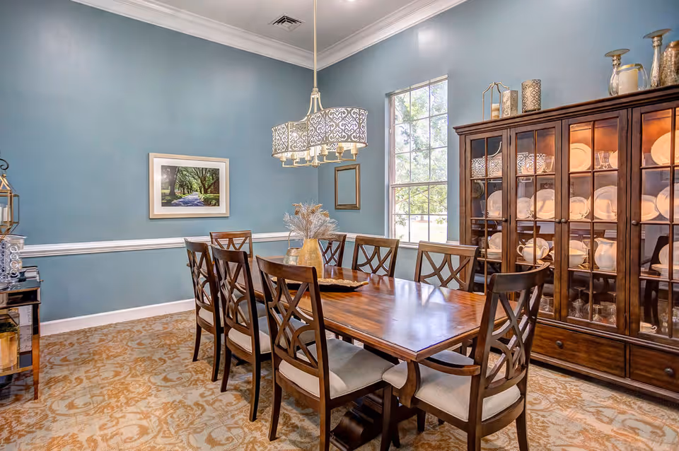 A dining room with a wooden table and eight matching chairs with cushioned seats. The walls are painted blue with white trim, and there is a large window letting in natural light. A chandelier with a decorative shade hangs above the table. On the right side, there is a wooden china cabinet filled with plates and glassware. A framed picture and a small mirror hang on the wall.