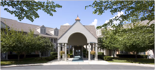 Front exterior view of Carrington Place Apartments featuring a covered entrance with columns, surrounded by trees and shrubs under a blue sky with some clouds.