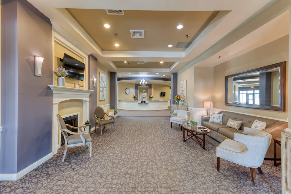 A spacious and well-lit senior living facility lobby with a patterned carpet, beige walls, and recessed ceiling lights. The area features a seating arrangement with a sofa, armchairs, a coffee table, and side tables with lamps. There is a fireplace with a mounted TV above it on the left side, decorative wall art, and a large mirror on the right wall. In the background, a reception desk with flowers and a clock on the wall is visible.