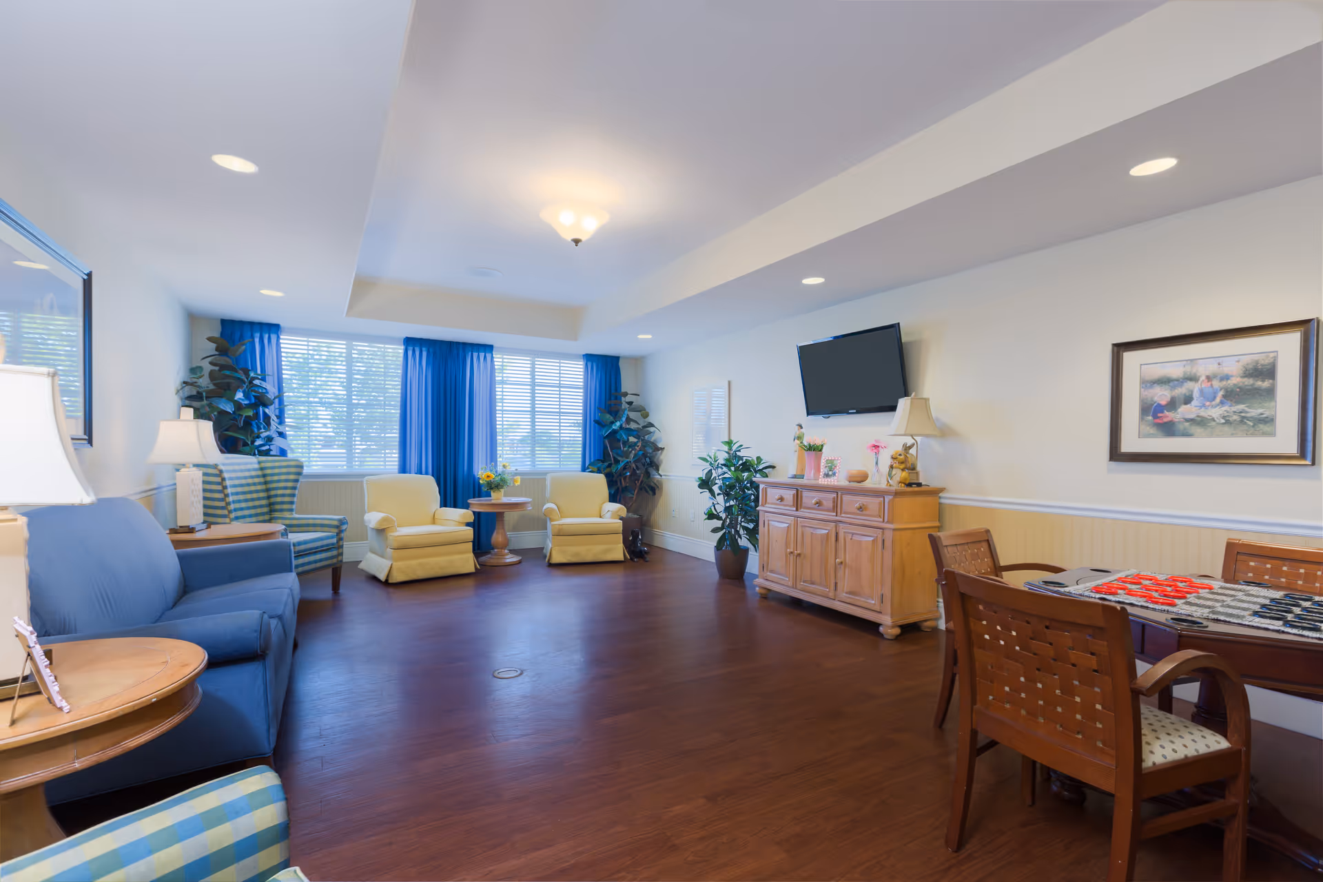 Bright communal living room with sofas and armchairs, a TV mounted above a wooden cabinet, and a table with chairs.
