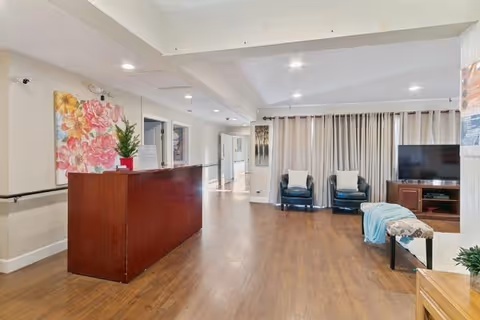 Interior view of a senior center reception area with a wooden reception desk on the left, a large floral painting on the wall behind it, and a hallway leading to other rooms. On the right side, there are two black armchairs, a TV on a wooden stand, and a bench with a blue blanket draped over it. The floor is wooden, and the ceiling has recessed lighting.