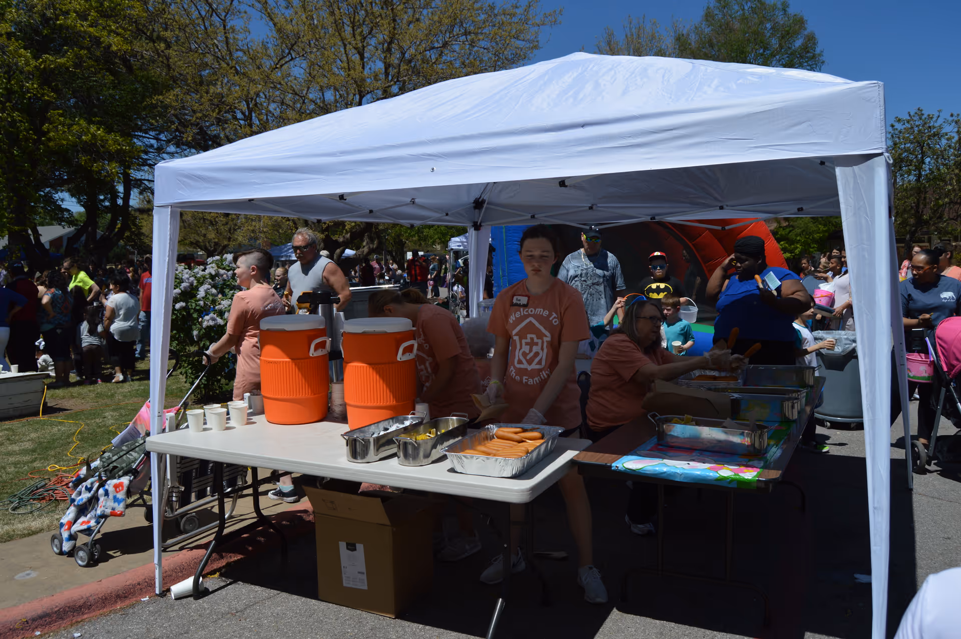 Outdoor event with a white canopy tent where volunteers in matching orange shirts are serving food and drinks to a crowd of people. Two large orange beverage coolers and trays of hot dogs are on tables under the tent. People, including children and adults, are gathered around the area enjoying the event on a sunny day with trees and blue sky in the background.