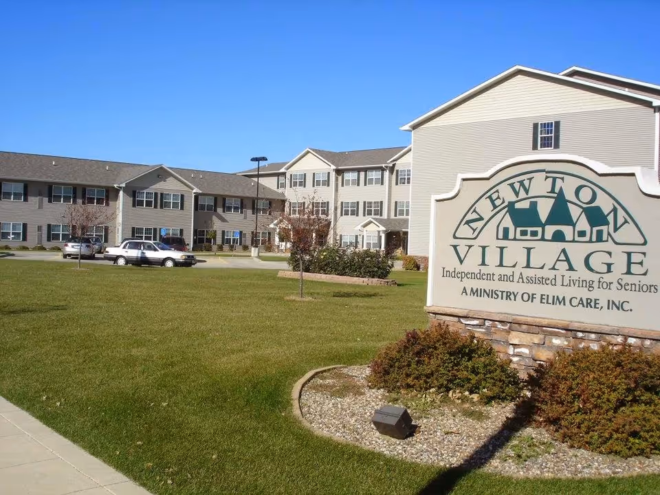 Exterior view of Newton Village, an independent and assisted living facility for seniors, showing a large beige building with multiple windows, a green lawn, a few parked cars, and a sign in the foreground with the facility's name and description.