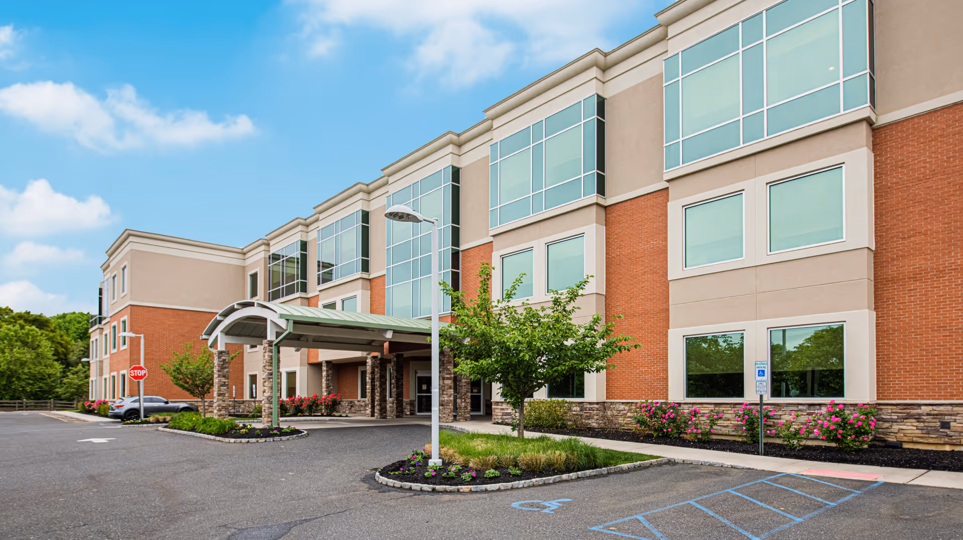 Front entrance of a modern three-story brick-and-stucco building with a covered drop-off, parking area, and landscaped beds.