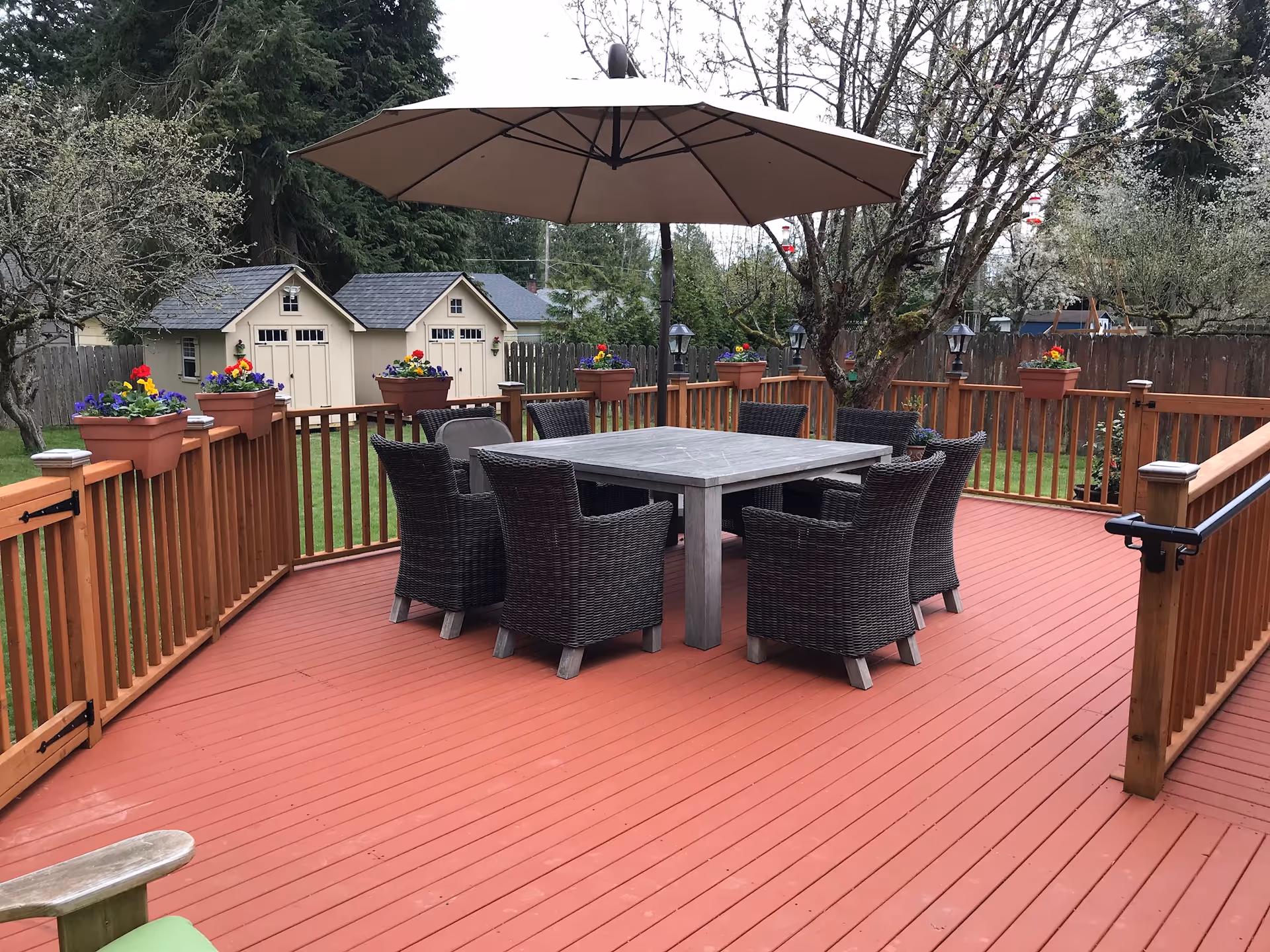 A spacious outdoor wooden deck with a square table surrounded by eight wicker chairs. A large beige umbrella is open above the table. The deck is decorated with flower pots containing colorful flowers attached to the wooden railing. In the background, there are two small beige garden sheds and trees with sparse leaves.