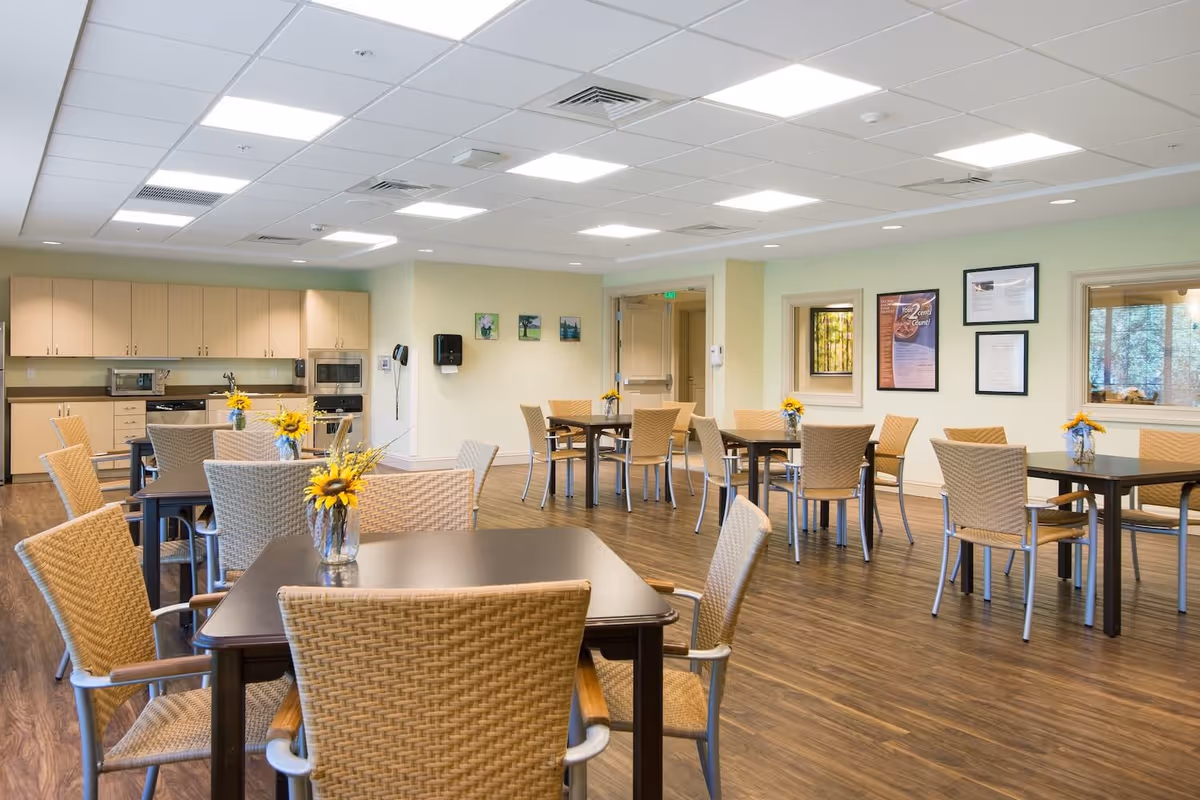 A bright and clean dining room with several dark wood tables and wicker chairs arranged neatly. Each table has a small vase with sunflowers. The room features wood flooring, light green walls, and a kitchen area with light wood cabinets, a microwave, and a dishwasher in the background. There are framed pictures and informational posters on the walls.