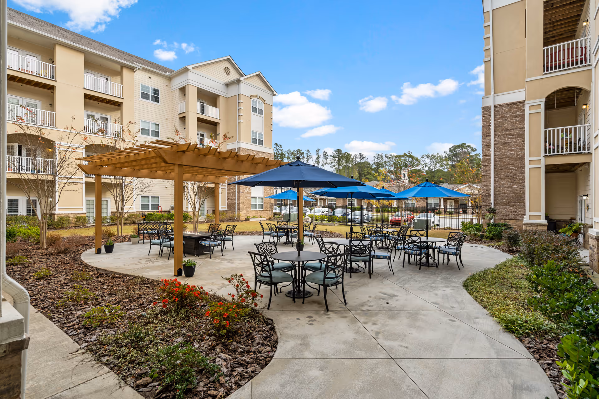 Outdoor patio area at a senior living facility with multiple round tables and chairs, some shaded by blue umbrellas. There is a wooden pergola with additional seating underneath. Surrounding the patio are landscaped garden beds with plants and flowers. In the background, there are multi-story residential buildings with balconies under a partly cloudy blue sky.
