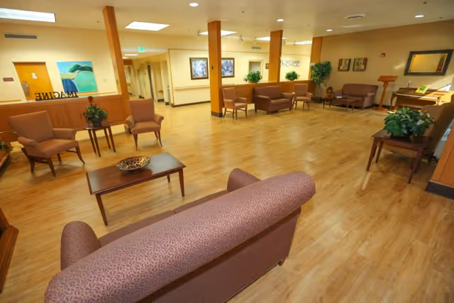 A spacious living room area with wooden flooring, multiple seating arrangements including sofas and armchairs, small tables with decorative plants, framed artwork on the walls, and a piano in the corner.