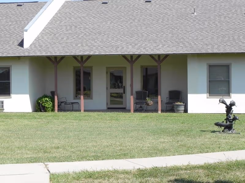 Front exterior of a single-story assisted living building with a covered patio, seating, lawn, and a small bird sculpture.