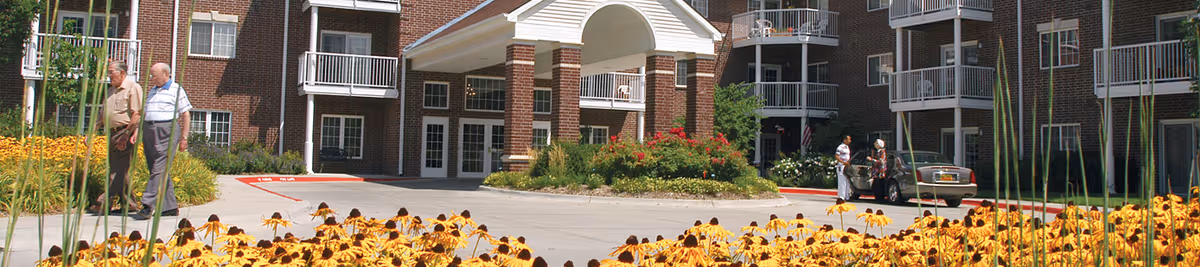 Front entrance of a brick retirement community with a porte-cochere, flower beds in the foreground, and residents walking nearby.