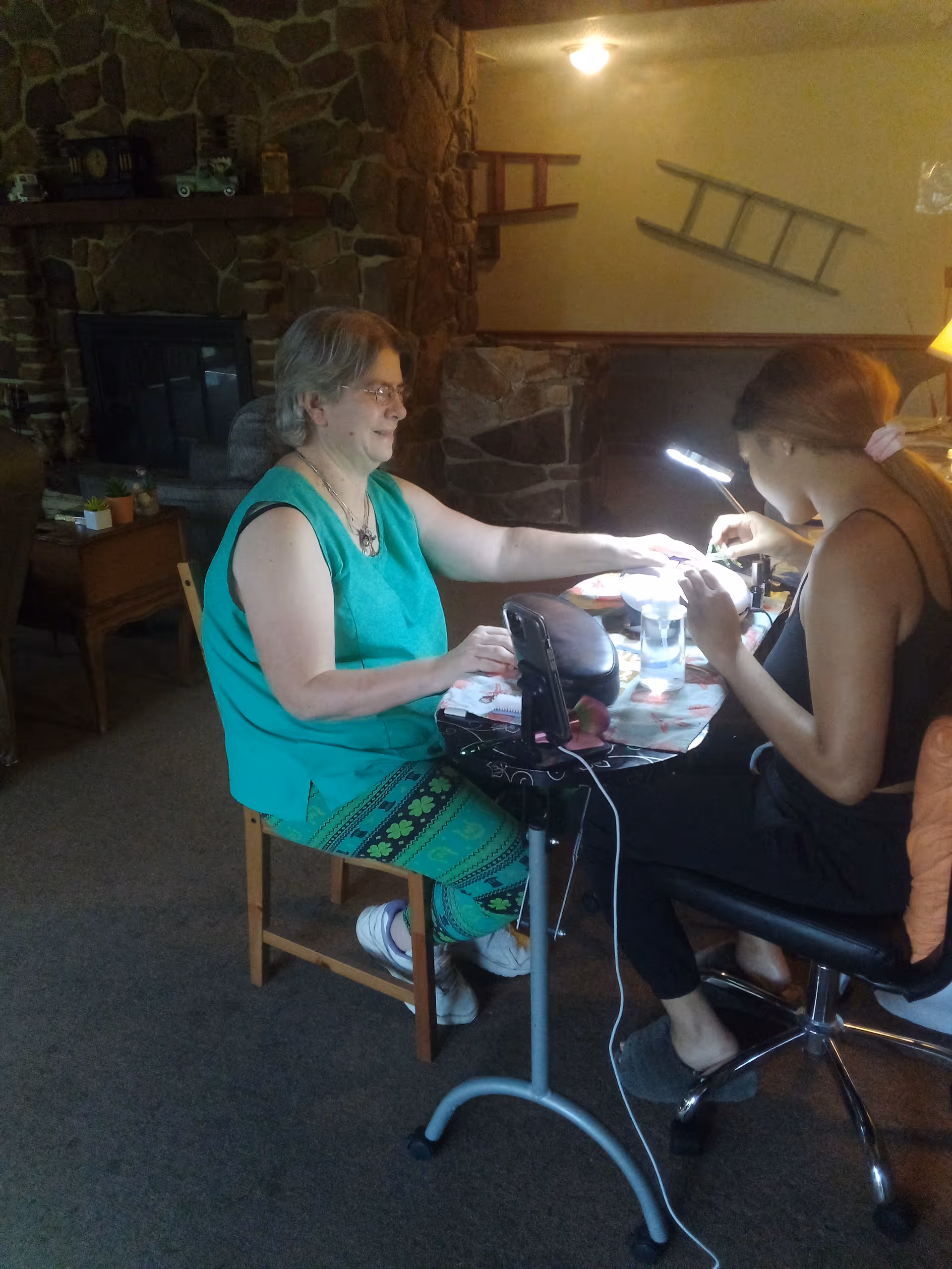 Two women sitting indoors near a stone fireplace. One woman with short gray hair and glasses is seated on a wooden chair wearing a green sleeveless top and patterned green pants. The other woman with long hair tied back is seated on a black swivel chair, focused on giving a manicure to the first woman. A small table with manicure tools and a lamp is between them. The room has a cozy atmosphere with stone walls and wooden furniture.