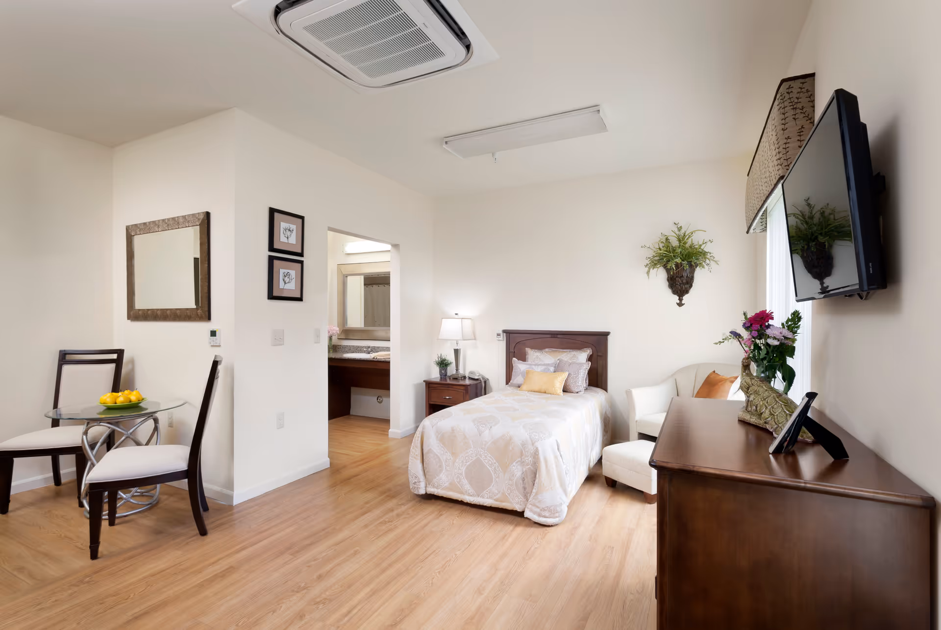 A bright, neatly arranged assisted living bedroom with a single bed covered in a patterned beige and white comforter. Next to the bed is a wooden nightstand with a lamp and a small plant. A cushioned armchair is placed near a window with a mounted flat-screen TV above a wooden dresser adorned with a vase of flowers and decorative items. On the left side of the room, there is a small round glass table with two chairs. The room has light wood flooring and cream-colored walls, with a doorway leading to a bathroom area visible in the background.