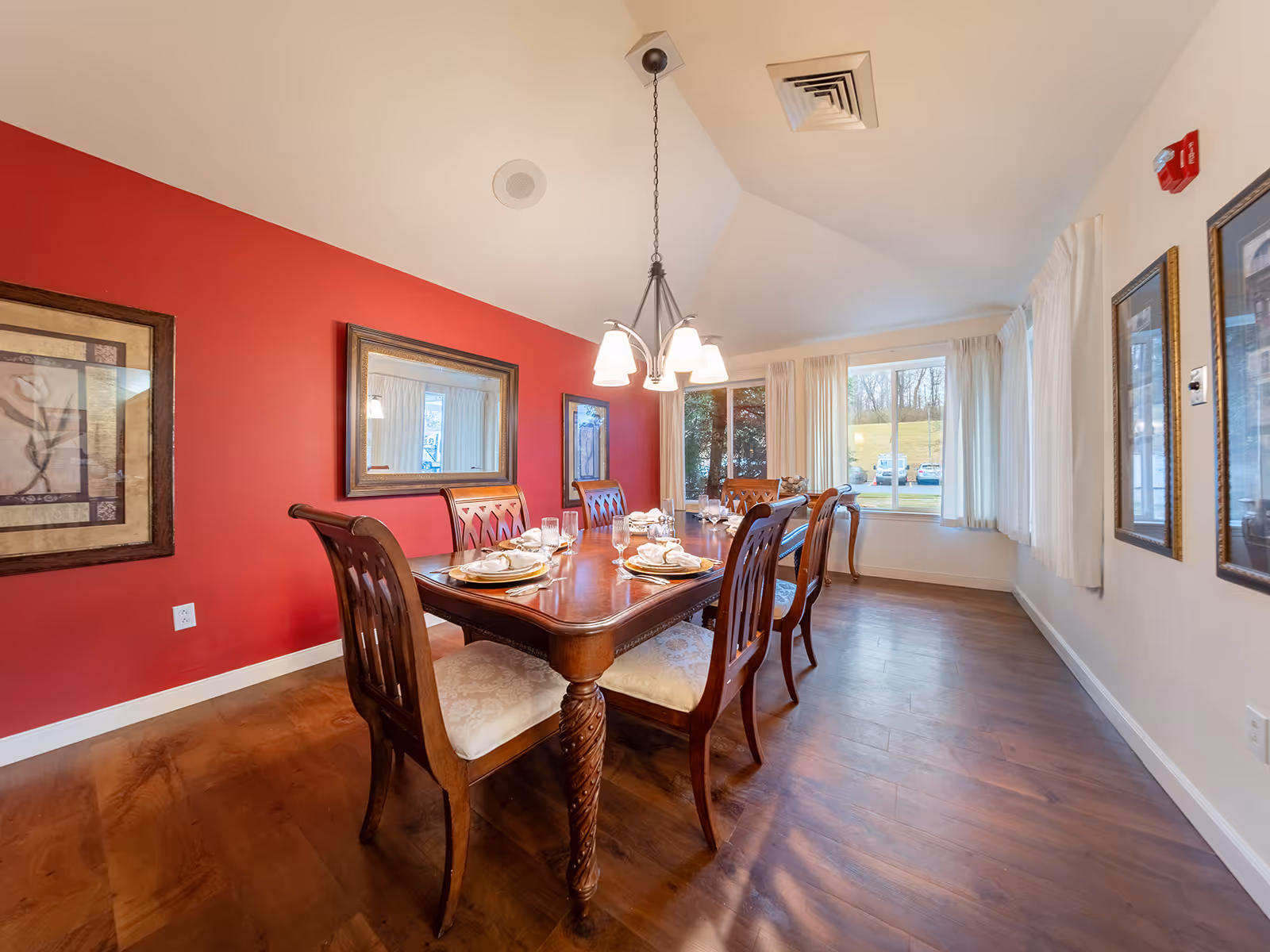 Formal dining room with a wooden table set for a meal, matching chairs, a red accent wall and a large window letting in daylight.