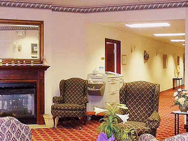 Cozy senior living common area with upholstered armchairs, a fireplace, plants, and a hallway.