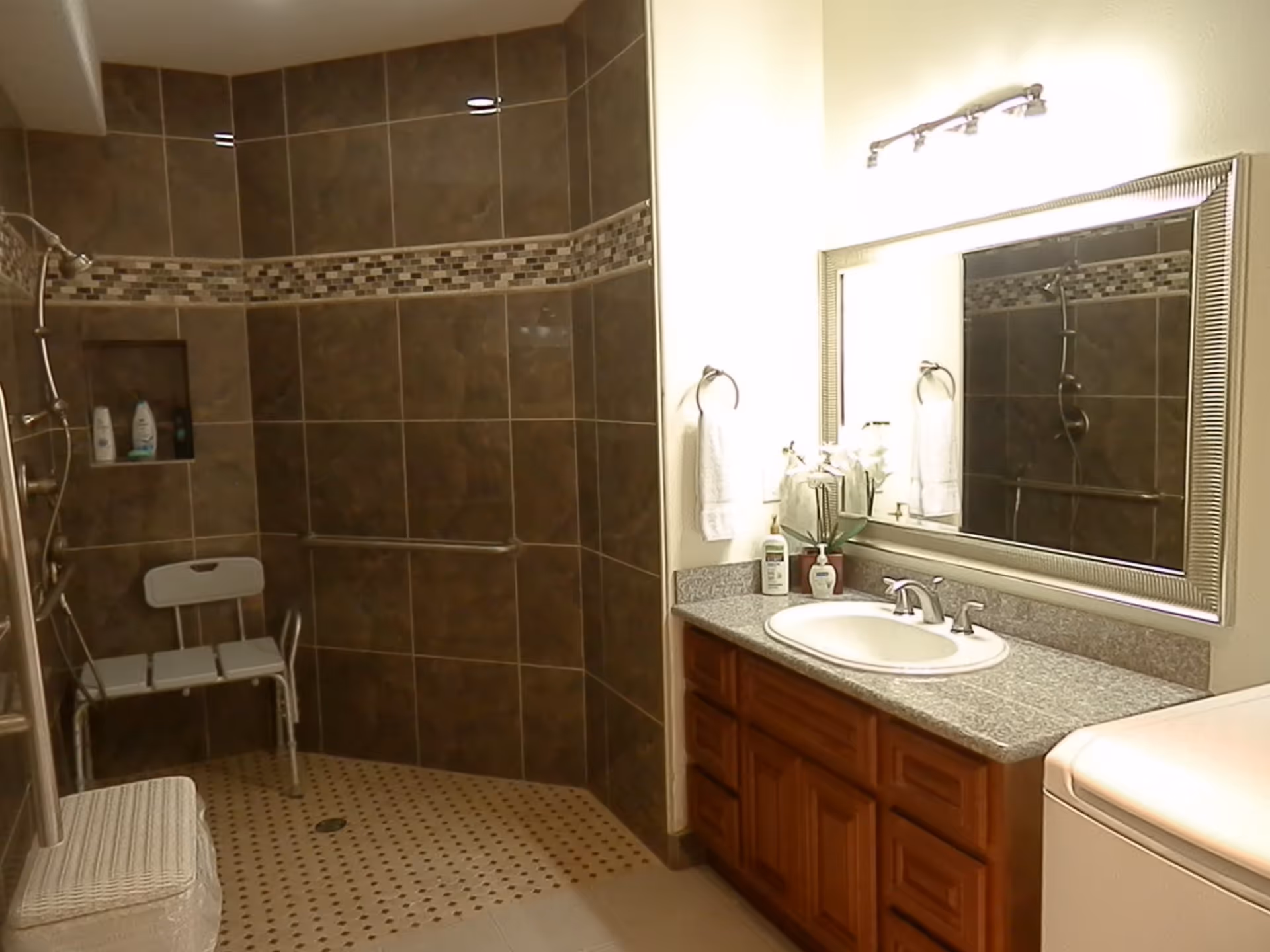 Bathroom with a walk-in shower featuring dark brown tiles and a mosaic tile accent strip. The shower has a built-in niche with toiletries and a white shower chair. There is a granite countertop with a sink, a large framed mirror, and a light fixture above. A towel ring with a white towel is mounted on the wall next to the sink. A white laundry basket is visible in the corner.