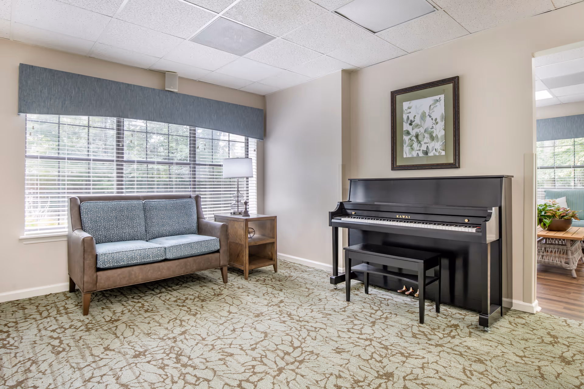 A bright common room with a black upright piano and bench opposite a small sofa and side table in front of large windows.