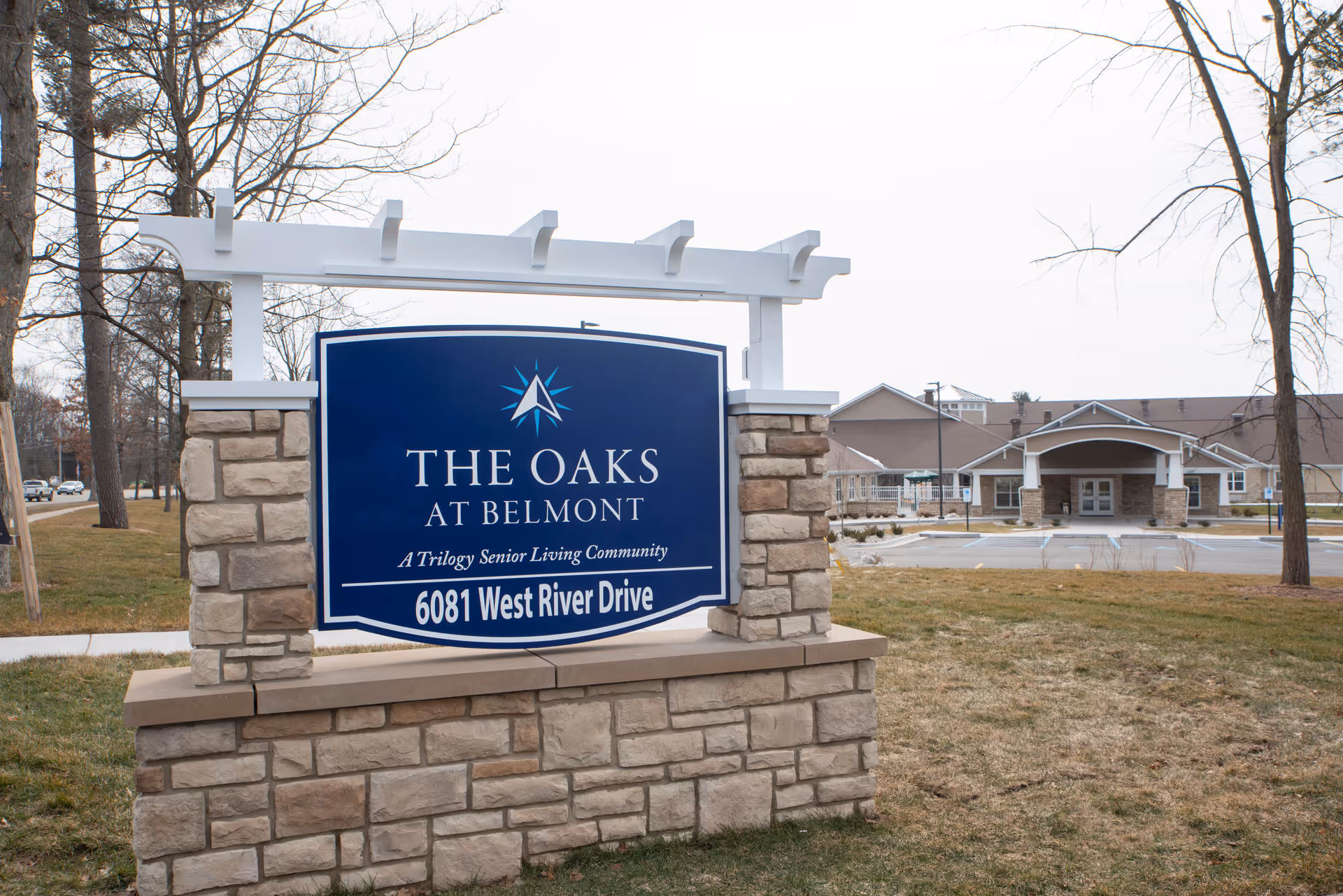 A large stone-and-wood entrance sign reading "The Oaks at Belmont" stands on a lawn with the senior living facility building visible in the background.