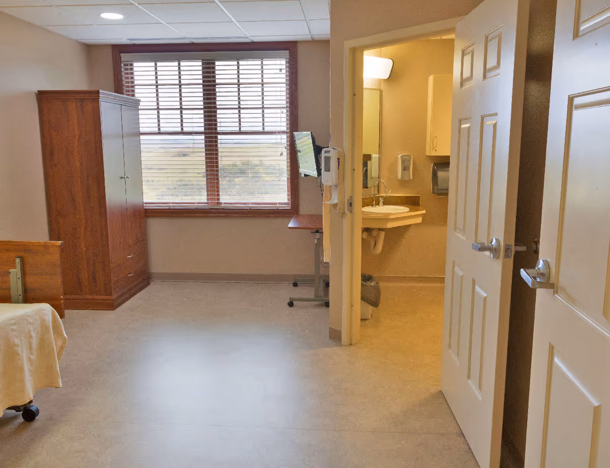 A room with a large window covered by wooden blinds, a wooden wardrobe, a hospital-style bed partially visible on the left, and an open door leading to a small bathroom with a sink, mirror, and wall-mounted soap dispenser.