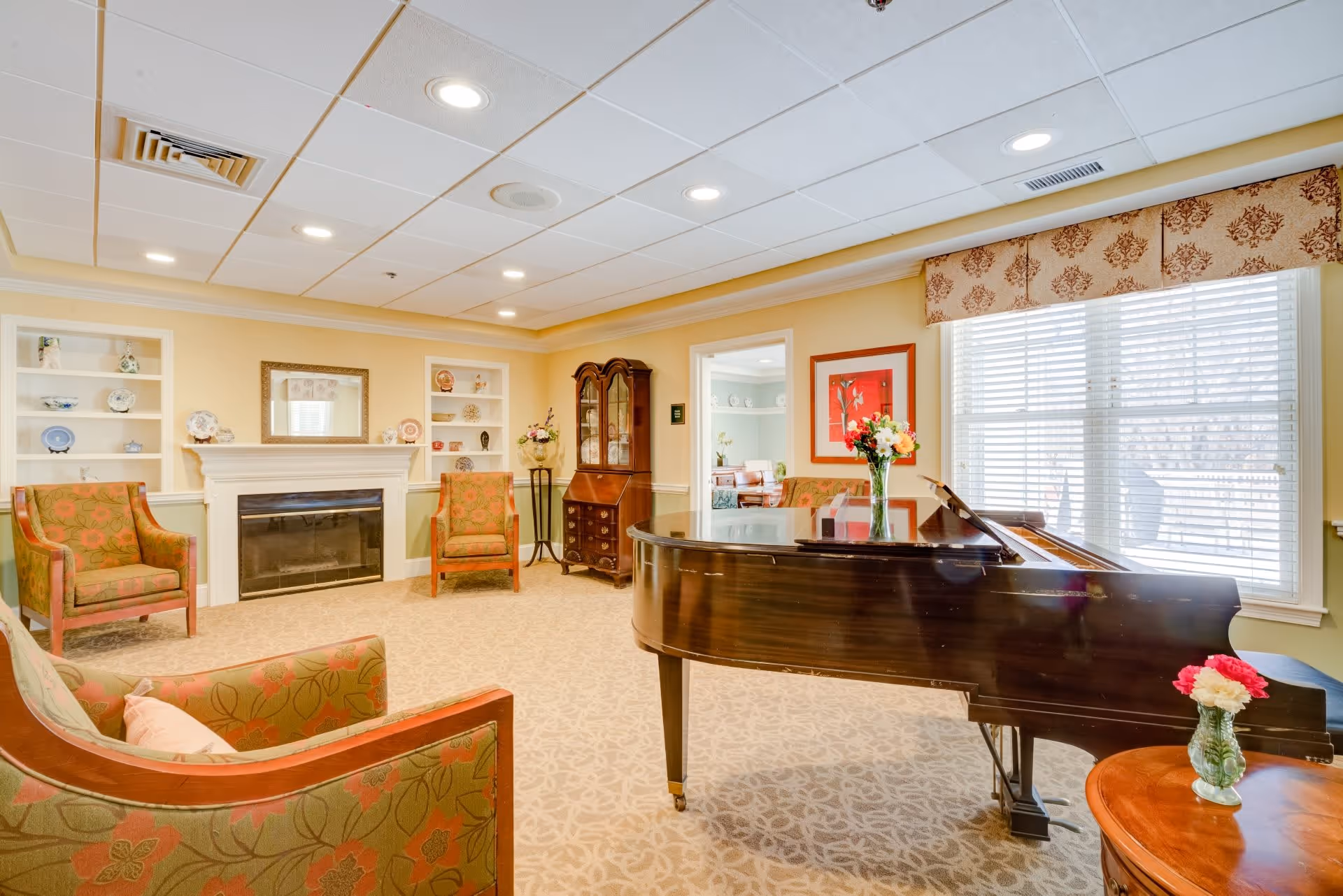 A cozy living room with a grand piano in the foreground, floral upholstered chairs, a fireplace with a mirror above it, built-in shelves with decorative plates, a wooden cabinet, and a large window with blinds and a patterned valance letting in natural light.