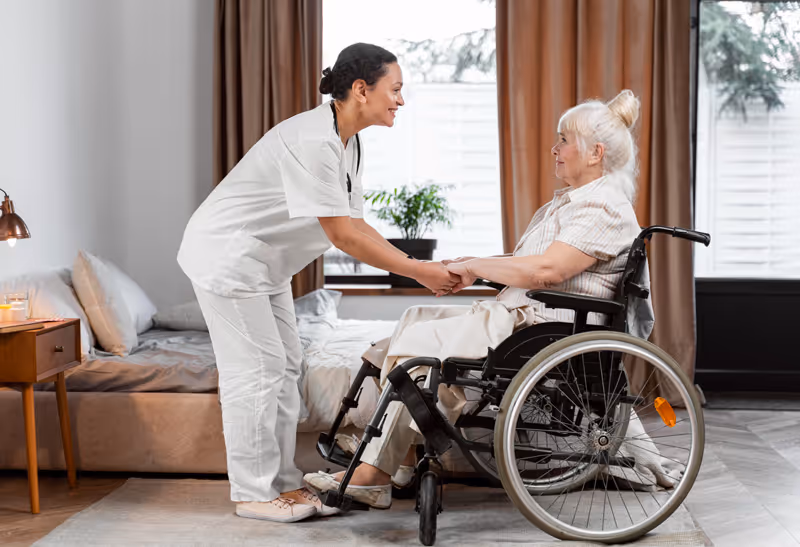 A caregiver interacts warmly with an elderly woman in a wheelchair inside a senior living facility.