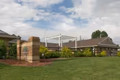 Exterior view of a single-story building with a brown roof and beige walls, surrounded by green grass, shrubs, and trees under a partly cloudy sky. There is a brick pillar structure in the foreground and a white pergola near the building.