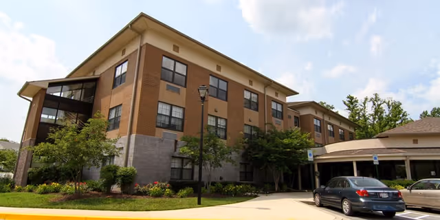 Exterior view of a three-story senior living facility building with brick and stone facade, surrounded by trees and shrubs, with a driveway and parked cars in front under a partly cloudy sky.