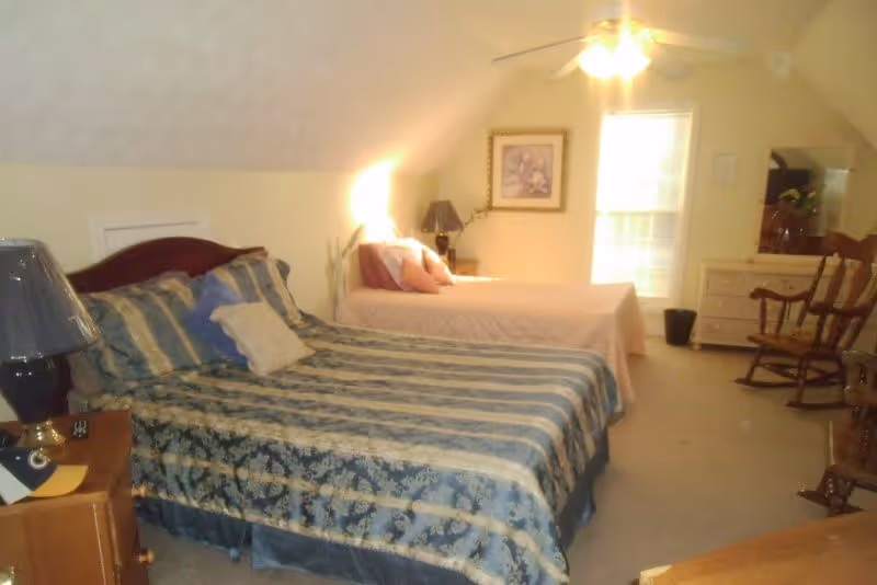 A bedroom with two beds, one with a blue and beige striped comforter and multiple pillows, and the other with a light pink comforter and pillows. There is a nightstand with a lamp between the beds, a framed picture on the wall, a window with blinds, a dresser with a mirror, and two wooden rocking chairs. The ceiling has a fan with lights.