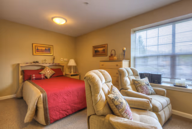 A cozy bedroom with a red-covered bed, two beige recliner chairs by a large window, a dresser and bedside table.
