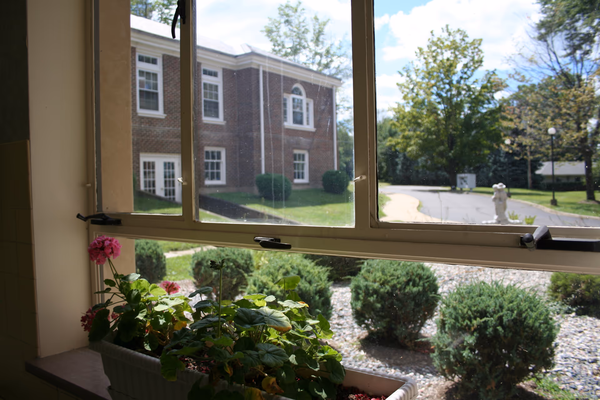 View through a window showing a flower box with pink and green plants on the windowsill. Outside, there are bushes, a paved pathway, trees, and a brick building under a partly cloudy sky.