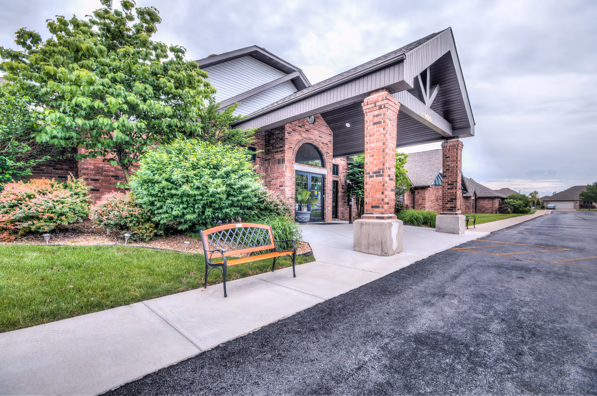 Entrance of a brick building with a covered driveway supported by brick columns, surrounded by green bushes and trees, with a bench on the sidewalk and a paved parking area to the right under a cloudy sky.