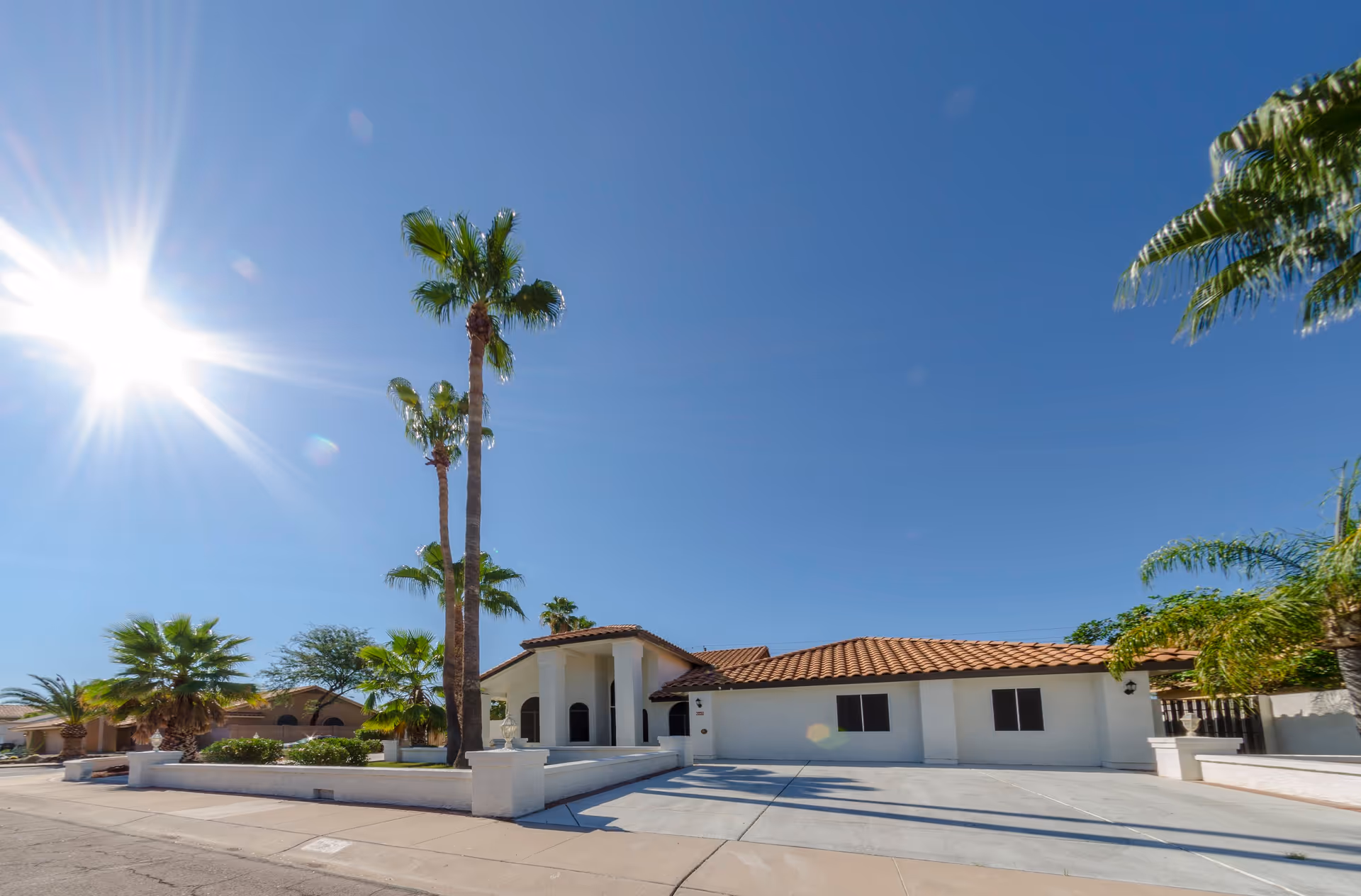 Single-story stucco house with a tiled roof, palm trees, and a wide driveway under a bright sun.