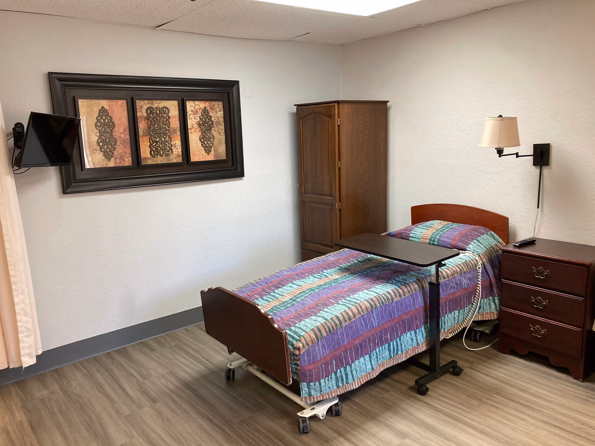 A small bedroom with a single hospital-style bed covered with a colorful striped blanket. Next to the bed is a dark wooden nightstand with three drawers and a wall-mounted lamp above it. A wooden cabinet is placed in the corner, and a flat-screen TV is mounted on the wall. Above the TV, there is a framed triptych artwork with decorative patterns. The floor is wood laminate, and the walls are light-colored.
