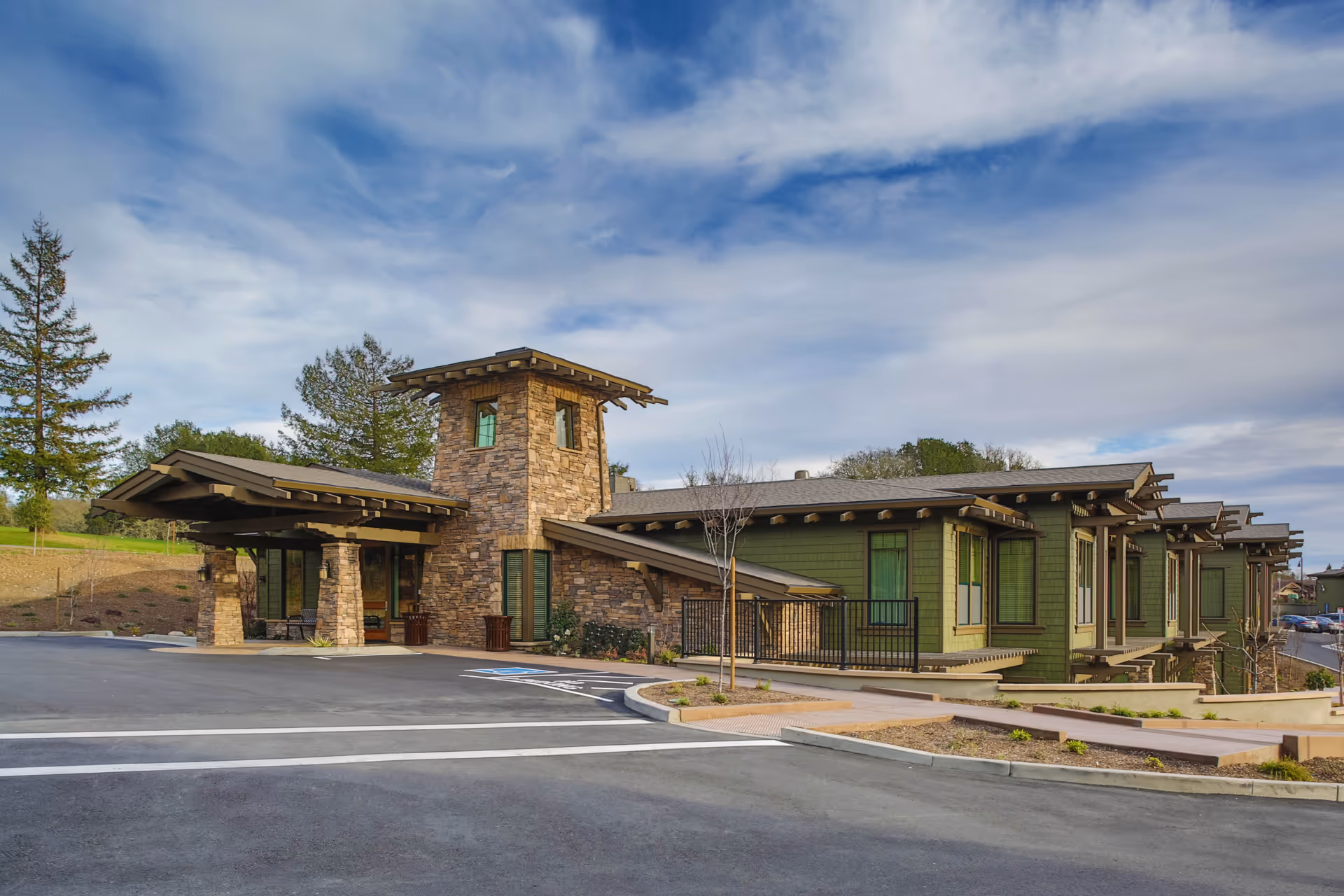 Exterior view of a single-story senior living facility building with green siding and stone accents, featuring a covered entrance and a tower-like structure. The building is surrounded by a paved parking area and landscaped grounds under a partly cloudy sky.