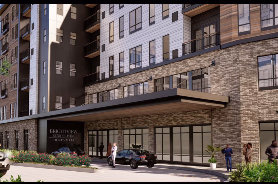 Exterior view of the Brightview Senior Living Hunt Valley building entrance with a covered drop-off area, several people standing and talking near parked cars, and landscaping with bushes and plants.
