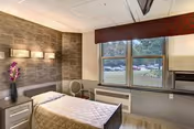 A single bedroom in a senior living facility featuring a bed with a beige quilted bedspread, a nightstand with a vase of purple flowers, a wall with modern lighting fixtures, a window with a view of parked cars outside, and a heating unit below the window.