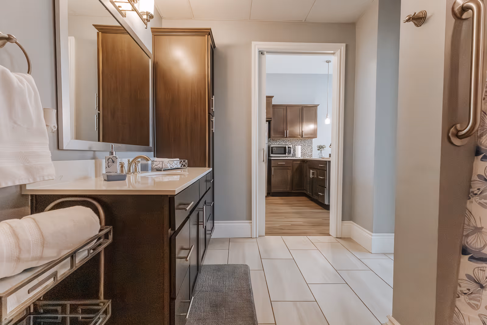 A modern bathroom with a large mirror above a dark wood vanity with a white countertop and a sink. White towels are neatly placed on a metal towel rack on the left. The bathroom floor is tiled in light colors. Through an open doorway, a kitchen with dark wood cabinets, a microwave, and a tiled backsplash is visible.