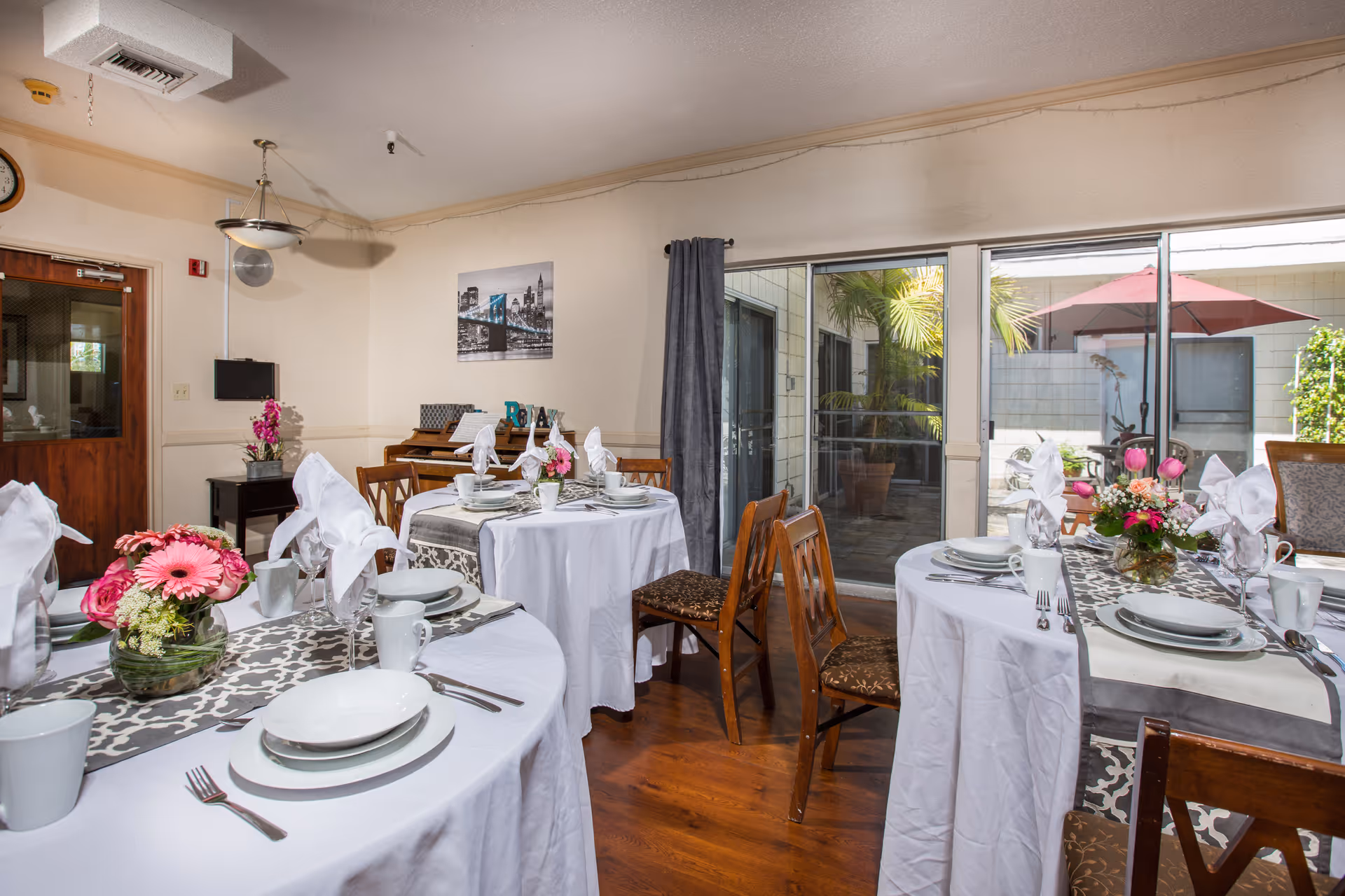 A dining room set up with round tables covered in white tablecloths, each table decorated with floral centerpieces and neatly arranged place settings including plates, cups, and folded napkins. The room has wooden chairs, a piano against the wall, a door leading outside, and large windows showing an outdoor patio with plants and a red umbrella.
