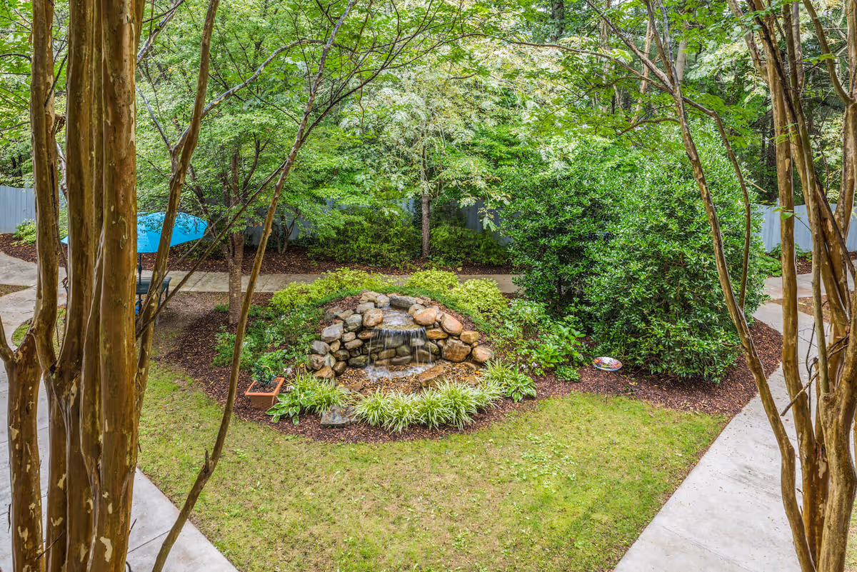 A peaceful outdoor garden area with a small rock waterfall feature surrounded by green bushes and trees. There are concrete pathways around the garden and a blue umbrella visible in the background.