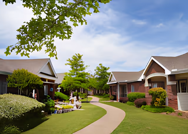 A winding paved pathway runs through a well-maintained garden area with green grass, bushes, and trees, flanked by single-story brick buildings with white trim under a partly cloudy blue sky.