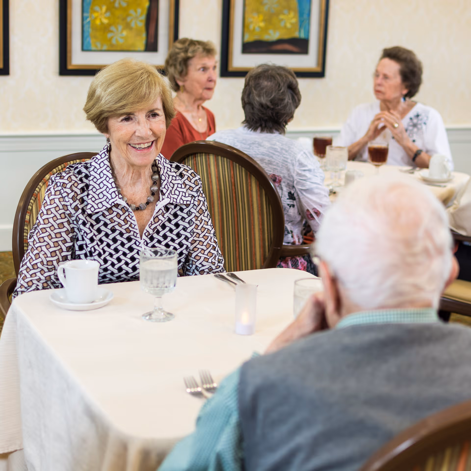 A smiling older woman sits at a table talking with other residents in a dining room.