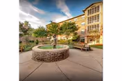 Courtyard with a circular fountain and statue, outdoor seating, and a multi-story senior living building in the background.