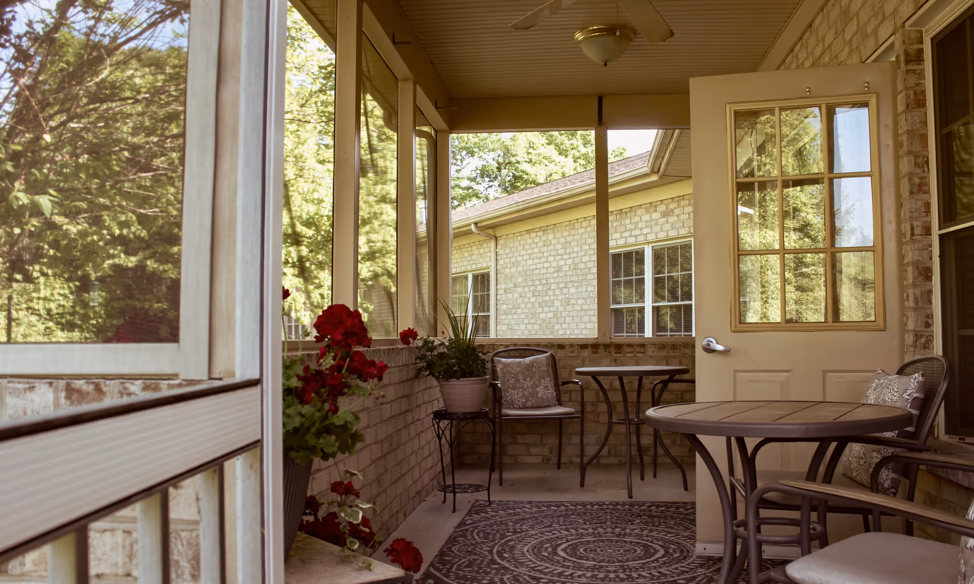 A cozy screened-in porch area with a round table, several chairs with cushions, potted plants, and a patterned rug. The porch is attached to a brick building and overlooks a green outdoor space with trees.