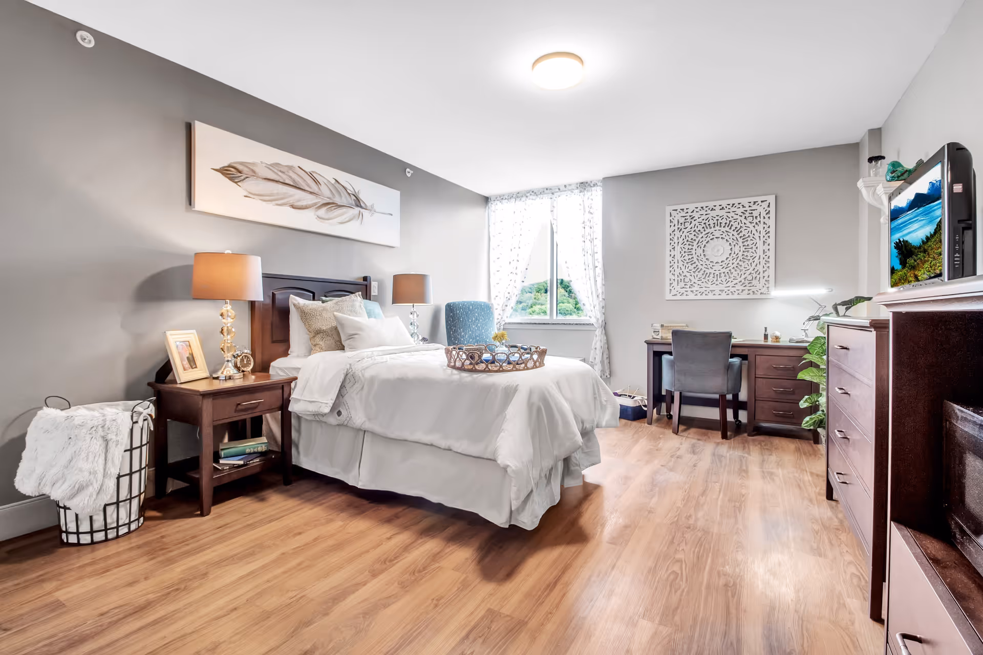 A bright and cozy bedroom with a single bed dressed in white linens and multiple pillows. The room features wooden flooring, a nightstand with a lamp, a framed photo, and an alarm clock. There is a blue armchair near a window with sheer white curtains letting in natural light. A wooden desk with a chair and a decorative wall piece is positioned against the far wall. A dresser with a TV on top is visible on the right side of the room.