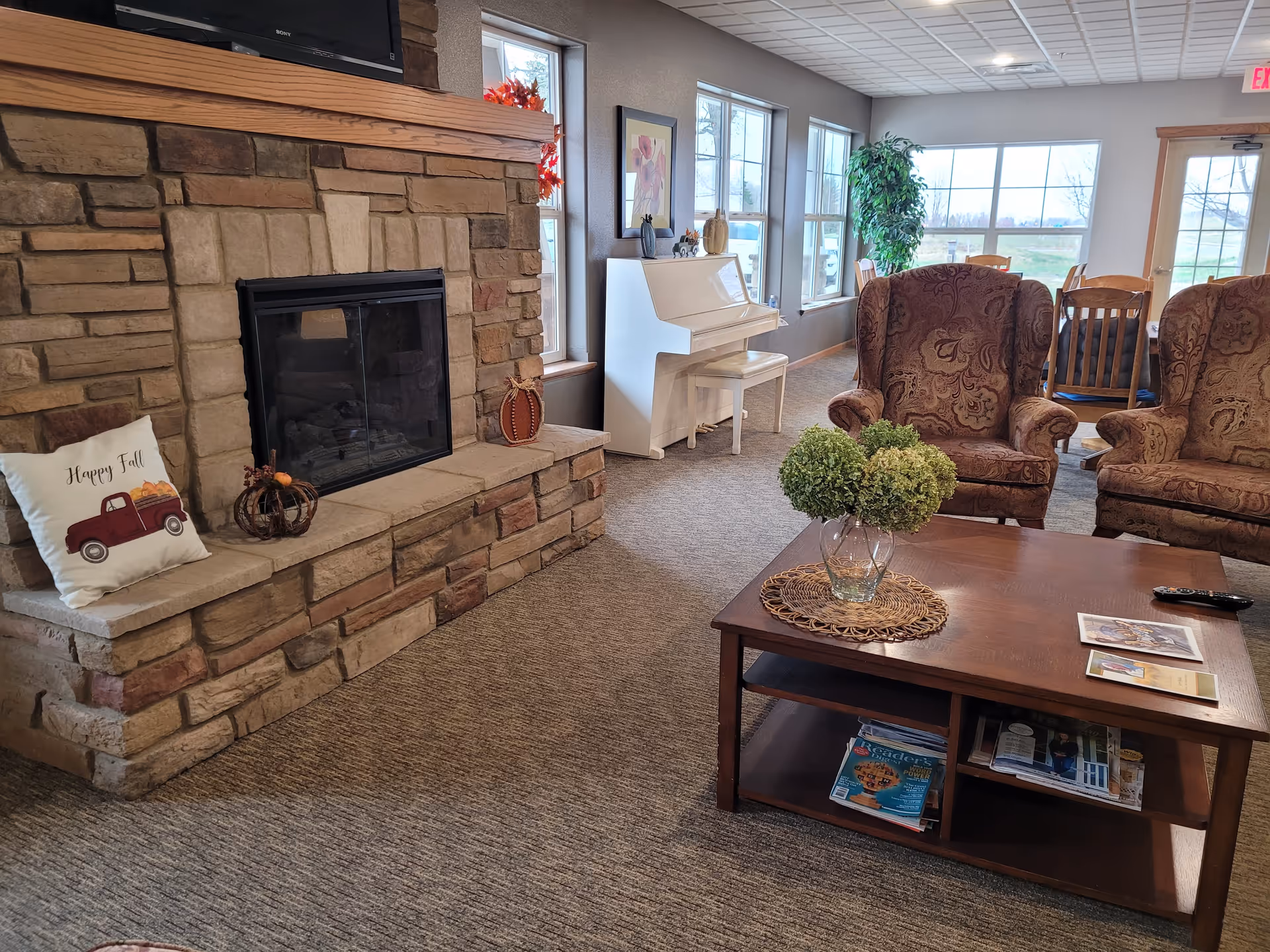 Cozy common room with a stone fireplace, a coffee table, upholstered armchairs, and a white piano by large windows.