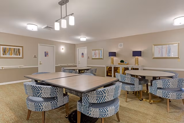 A well-lit interior room with multiple tables and patterned chairs arranged around them. The walls are beige with framed abstract artwork and two table lamps with dark blue shades on a sideboard. The room has a carpeted floor and ceiling lights.