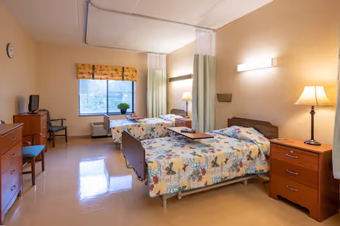A bright shared nursing facility bedroom with two beds with floral bedspreads, bedside tables and lamps, privacy curtains, and a window.