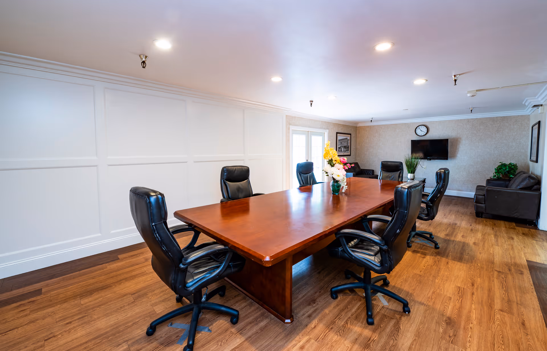Interior meeting room with a long wooden conference table surrounded by black leather chairs, a vase of flowers, a wall-mounted TV and a seating area.