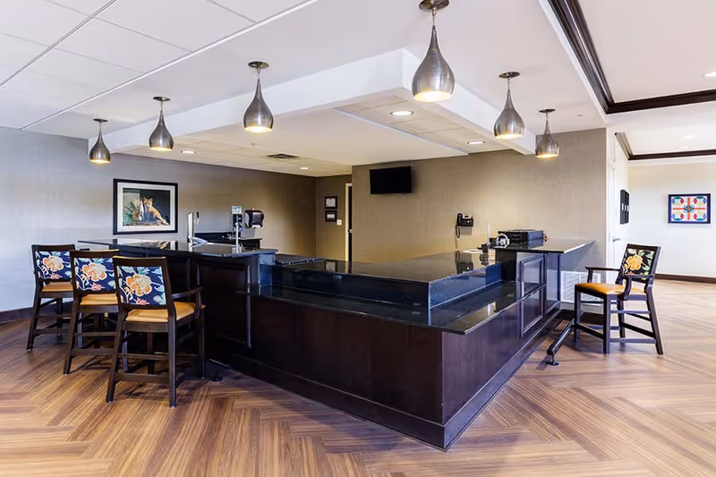 Interior view of a reception or common area with a large dark wood counter and black granite top. There are five wooden chairs with floral patterned cushions around the counter. The room has wood-patterned flooring, beige walls, and several pendant lights hanging from the ceiling. A framed picture and a wall-mounted TV are visible in the background.