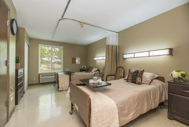 A well-lit senior care facility room with two single beds separated by a privacy curtain. Each bed has beige bedding and decorative pillows. There are bedside tables with flowers, chairs, and a window with blinds letting in natural light. The room has a clean and calm atmosphere.